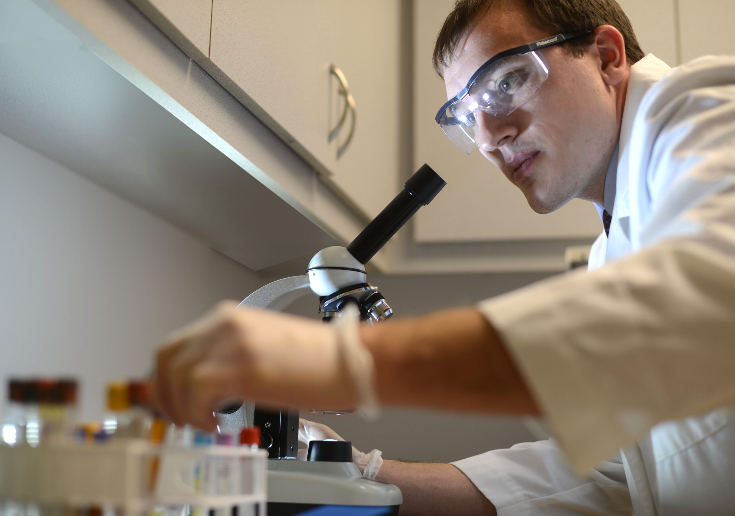 person wearing lab coat and safety glasses, holding test tubes and looking at microscope